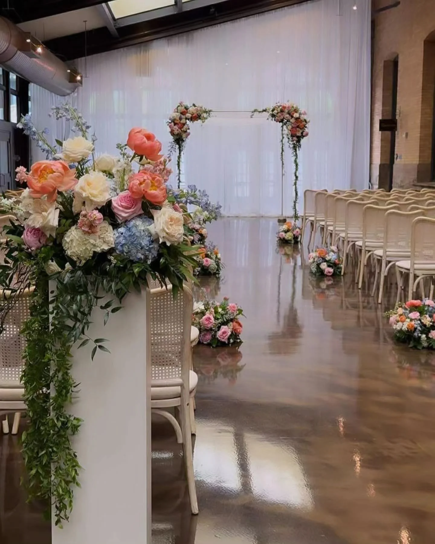 Indoor wedding ceremony setup with floral arrangements on a pedestal and along the aisle, and a floral arch at the front, in a room with wooden floors and white curtains.