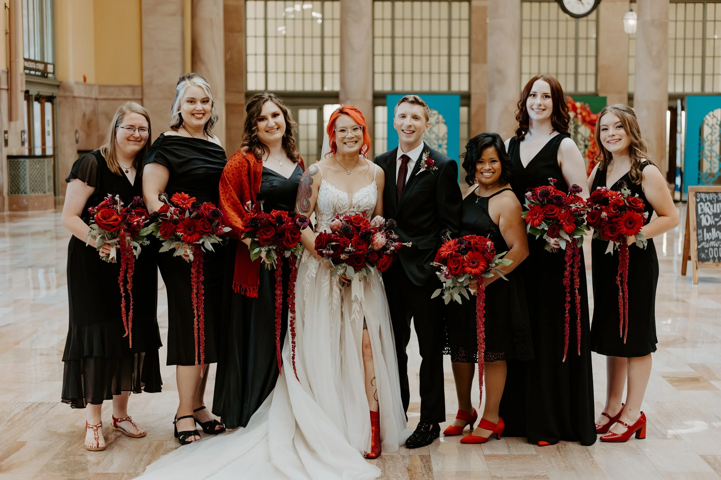 A group of nine people, including the bride and groom, in formal attire at a wedding. They are standing indoors in a spacious building with large windows and marble floors, holding bouquets of red and purple flowers.