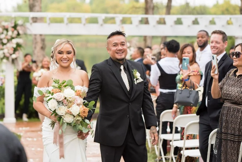 A bride and groom walking down the aisle at their outdoor wedding, smiling, with guests capturing the moment.