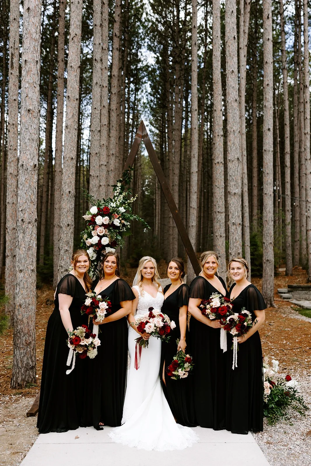 A bride and five bridesmaids smiling and holding bouquets during a wedding ceremony outdoors in a forest, with a floral arch and a triangular wooden frame in the background.
