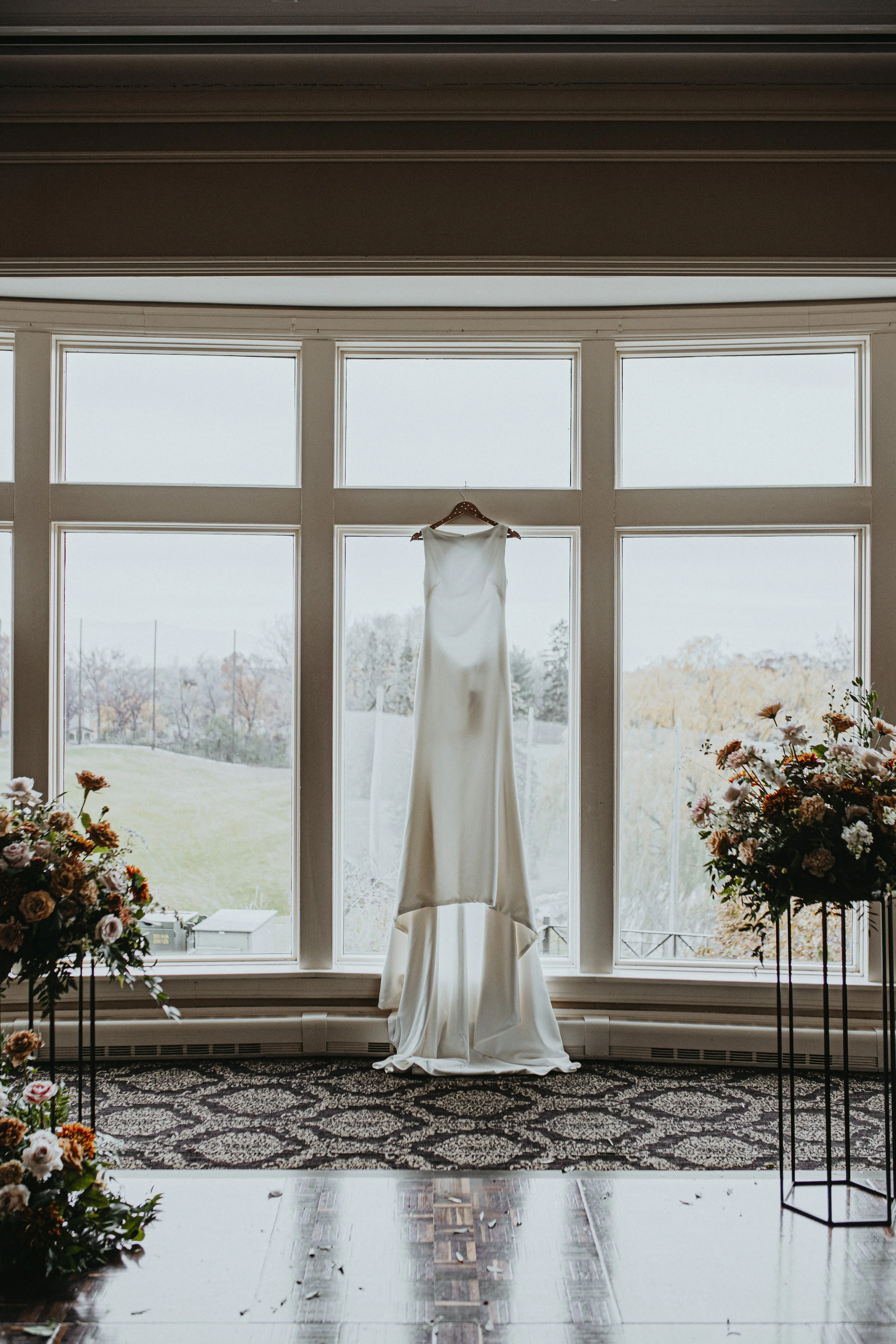 A white wedding dress hangs from a hook in front of a large window, with a view of a cloudy outdoor landscape and trees with fall foliage. Floral arrangements are visible on either side of the window.