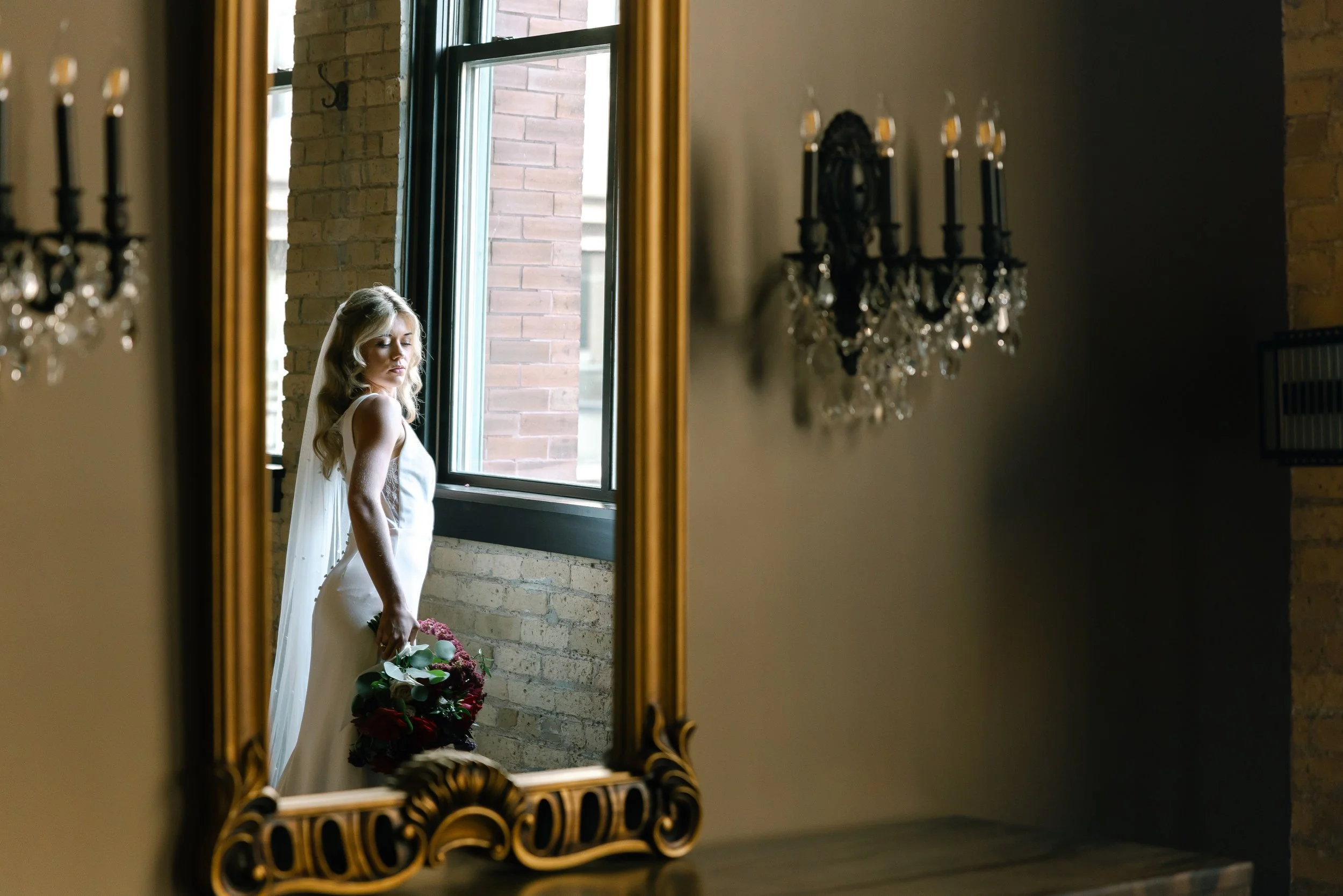 A bride standing in front of a window, holding a bouquet, as seen through an ornate gold-framed mirror.