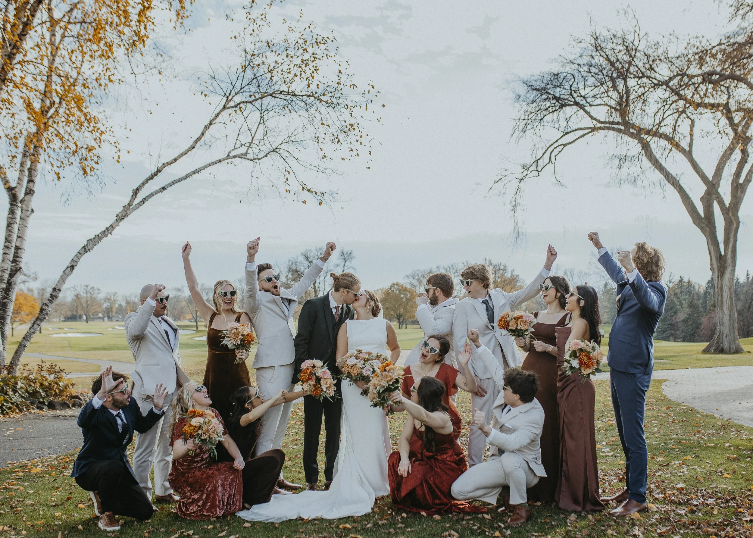 Group of people at a wedding outdoors, some standing and others kneeling on grass, with trees in the background. The bride and groom are in the center, kissing, surrounded by friends and family celebrating cheerfully.