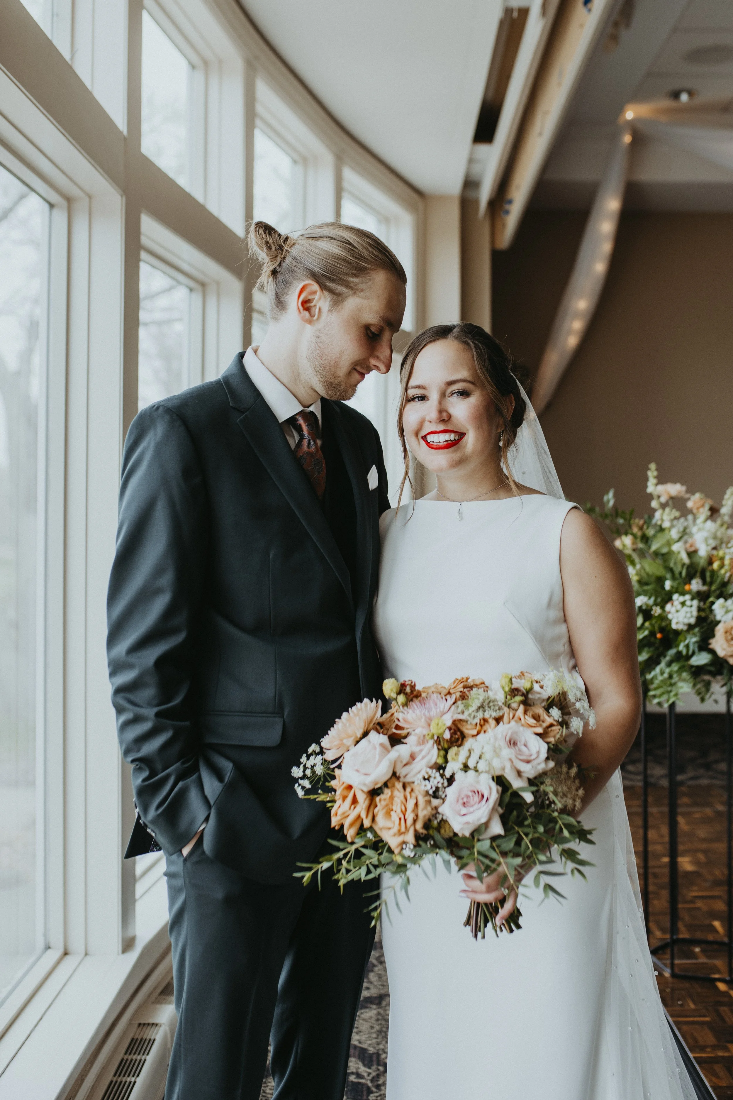 A bride and groom stand near large windows in an indoor setting, with the bride holding a bouquet of flowers and smiling at the camera, while the groom looks down with a gentle expression.