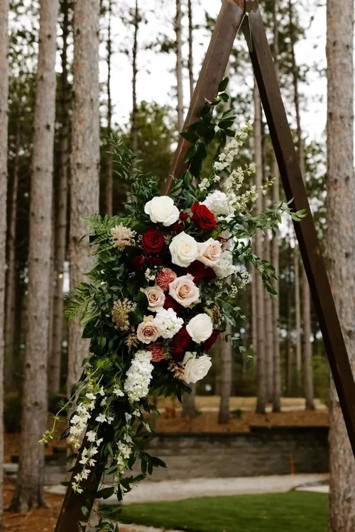 Flower arrangement with red and white roses, white lilies, and greenery on a wooden A-frame in a forest setting.