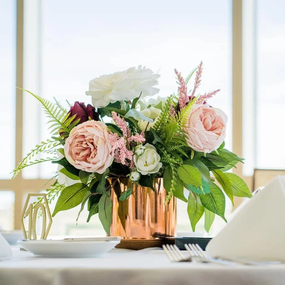 A pink metallic vase holding a bouquet of pink roses, white flowers, and greenery on a table set for a meal.