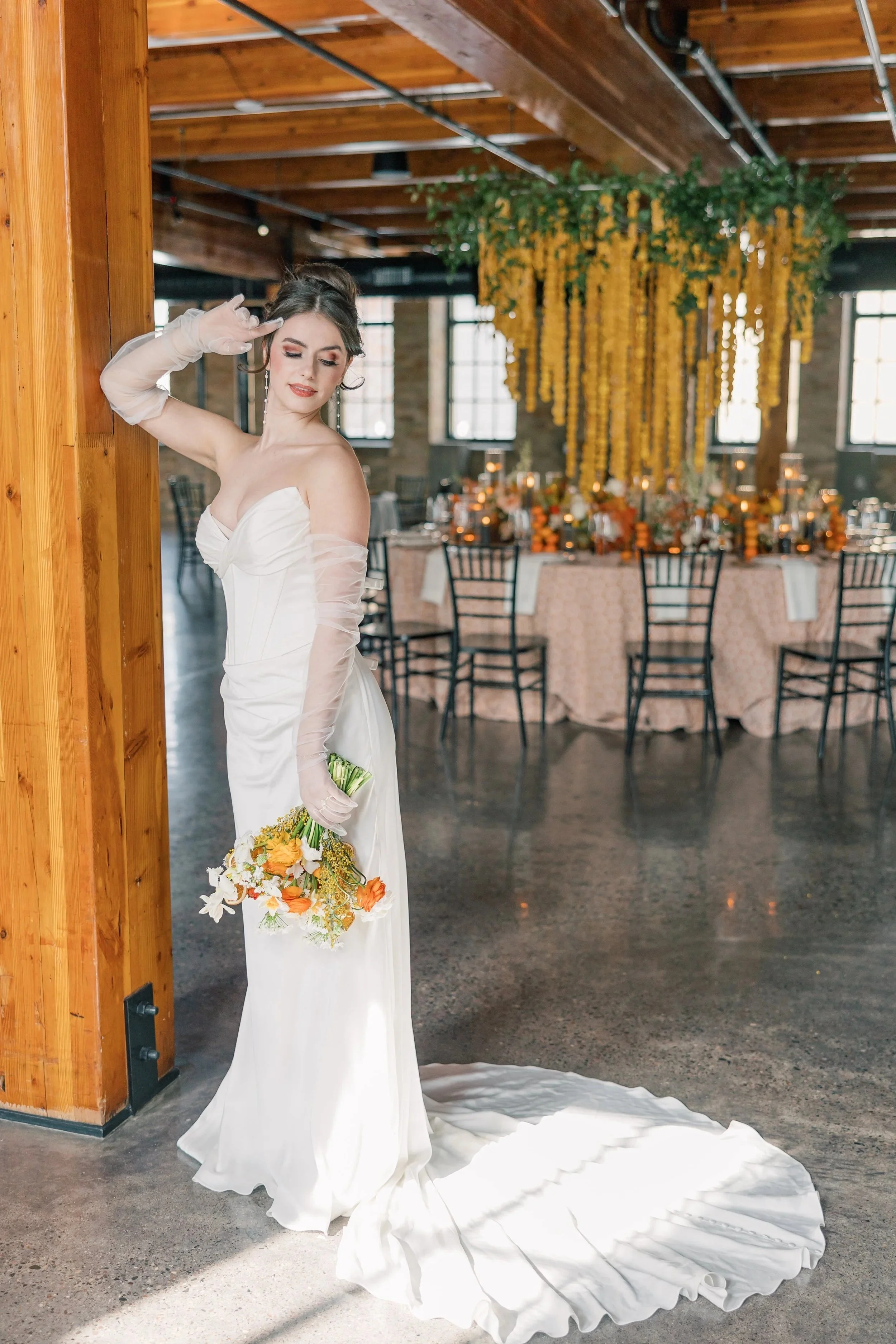 A bride in a strapless white wedding dress is holding a bouquet of orange and white flowers. She is standing in a rustic venue with wooden beams and large windows, with a floral and candle decorated table in the background. The bride is wearing long gloves and has her hair styled up, with makeup and earrings.