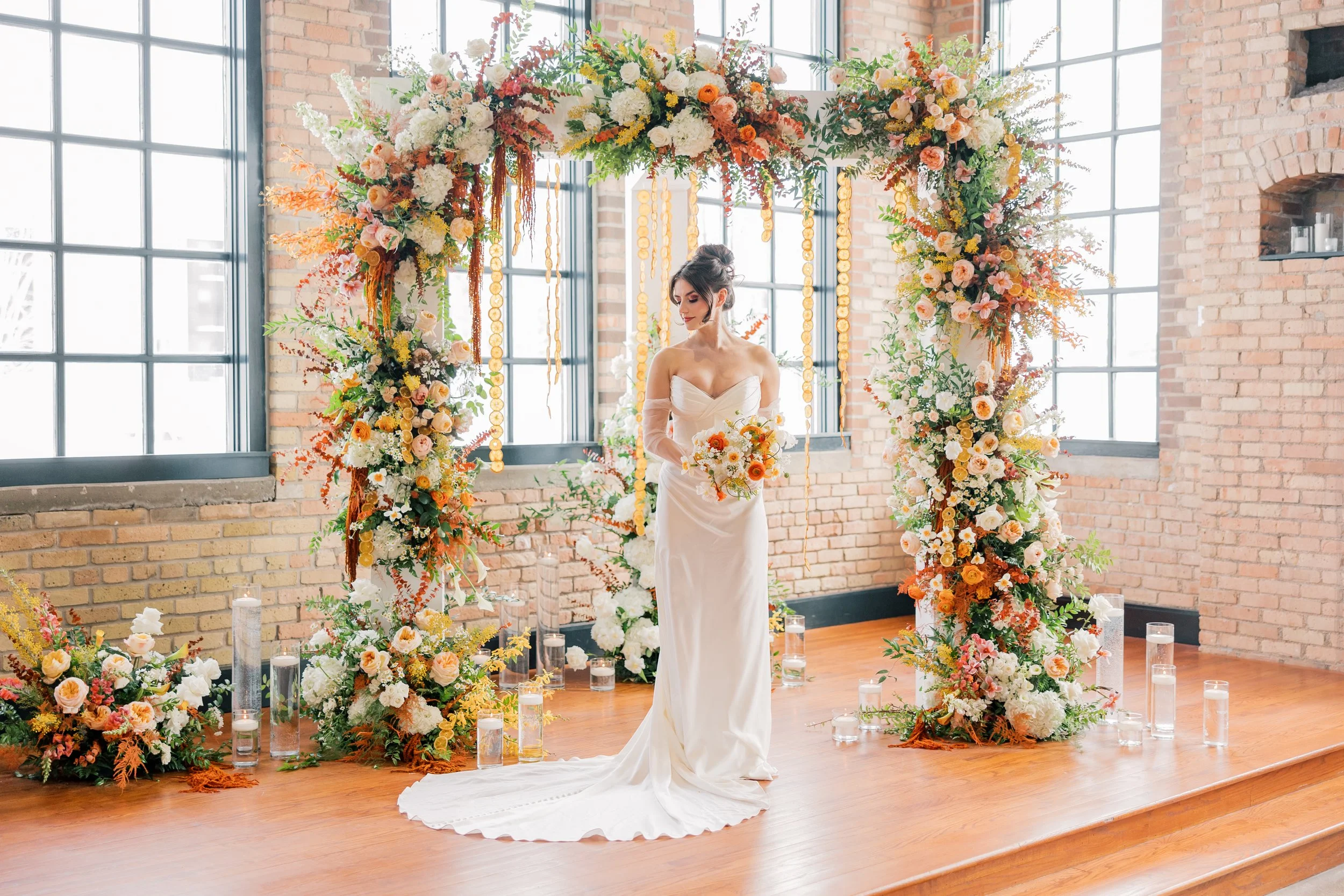 A woman in a white wedding dress stands beneath a colorful floral wedding arch in a bright, industrial-style venue with brick walls and large windows, surrounded by floral arrangements and candles.