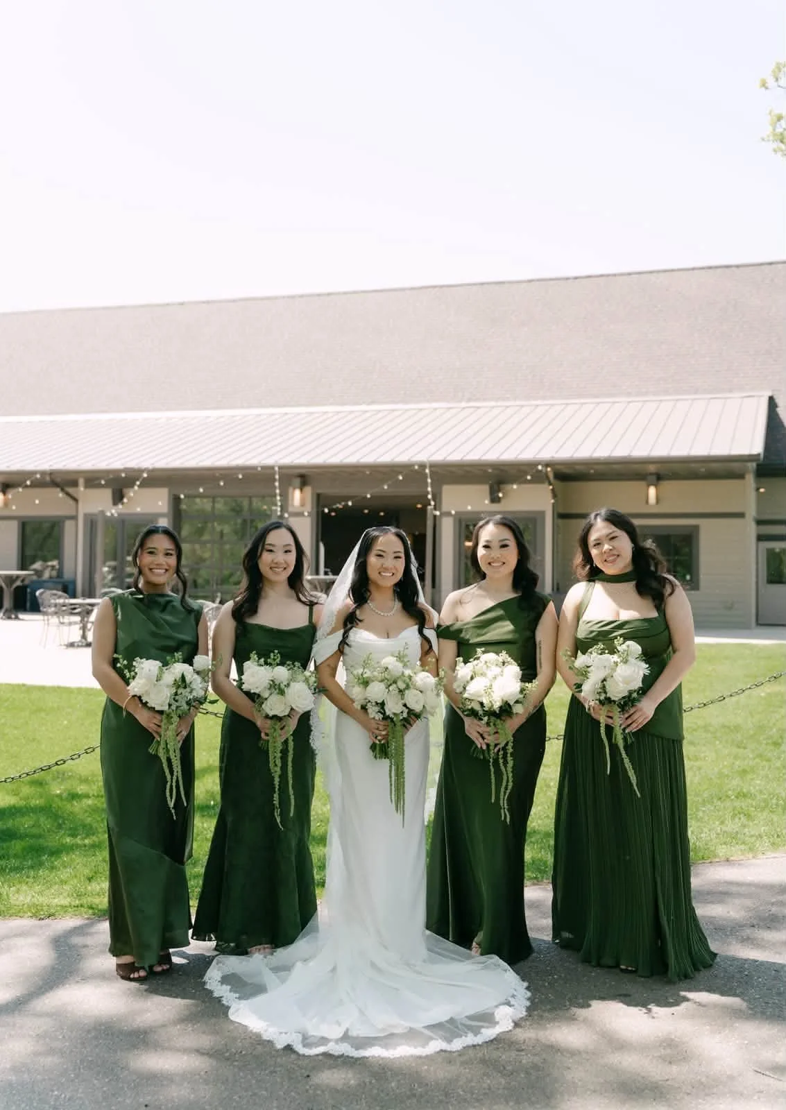 A bride in a white wedding dress and five bridesmaids in dark green dresses standing outdoors in front of a building with string lights, each holding a bouquet of white flowers.