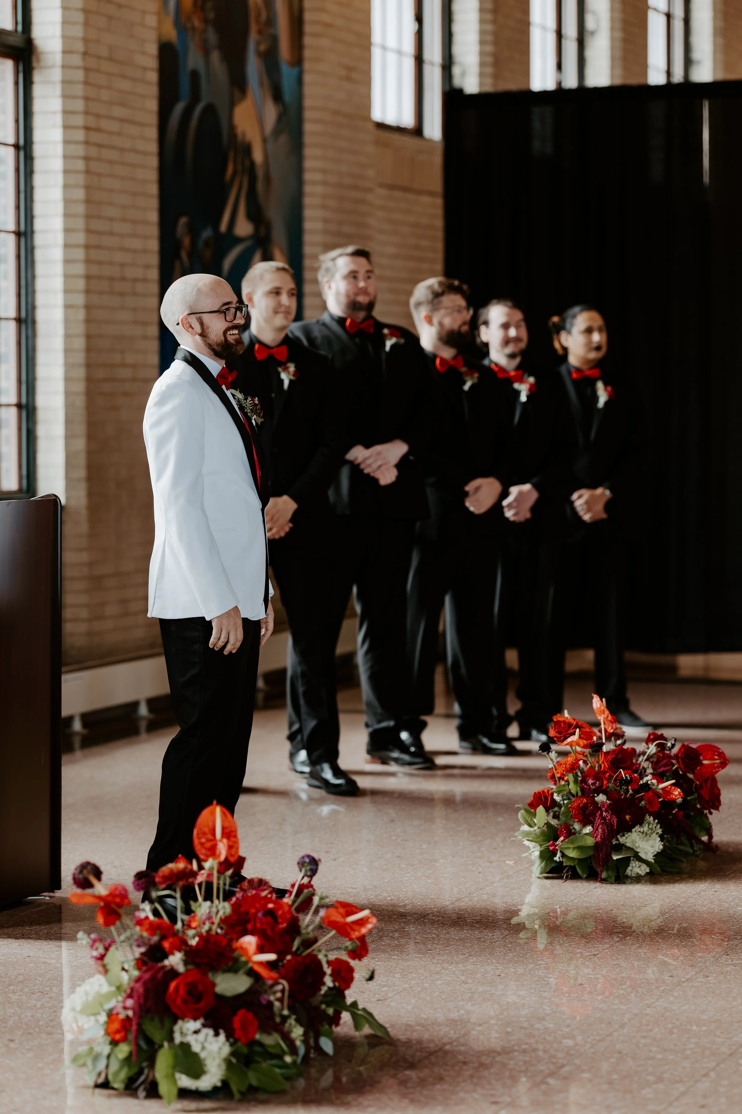 A groom in a white tuxedo with a black tie and red bowtie stands with a group of groomsmen in black suits, red bowties, and red floral boutonnieres inside a church, near colorful flower arrangements, during a wedding ceremony.