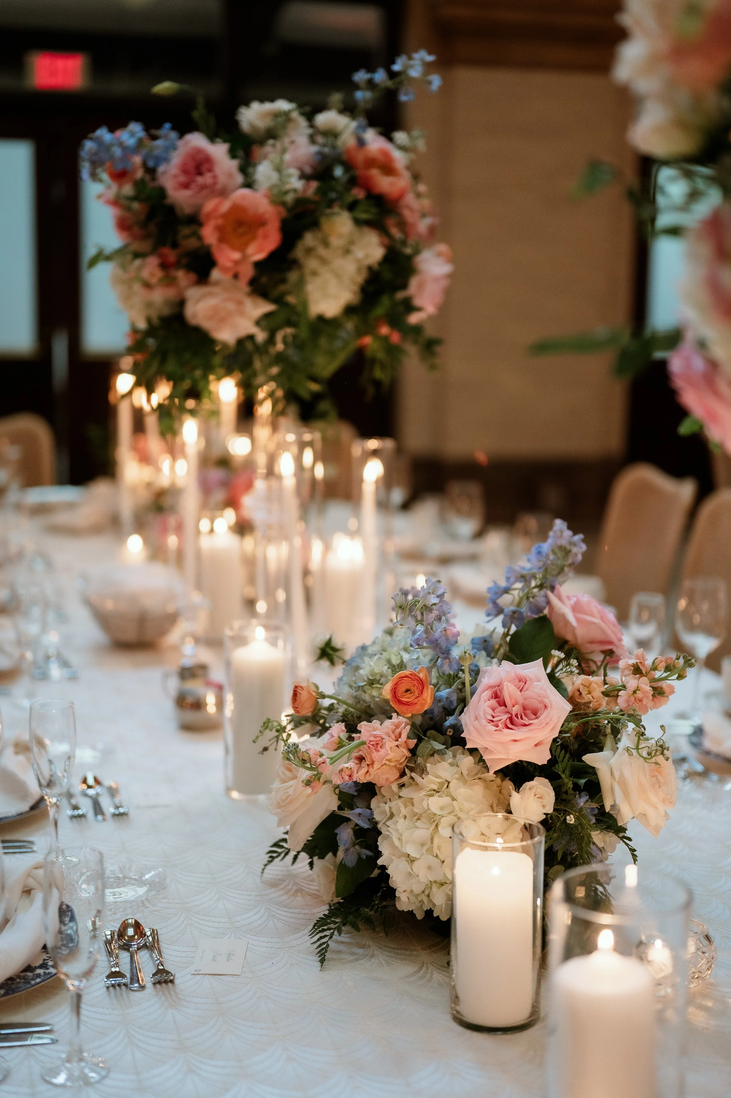 A close-up of an elegant table centerpiece with a large bouquet of pink, white, and purple flowers, surrounded by lit white candles in glass holders at a formal event or wedding reception.