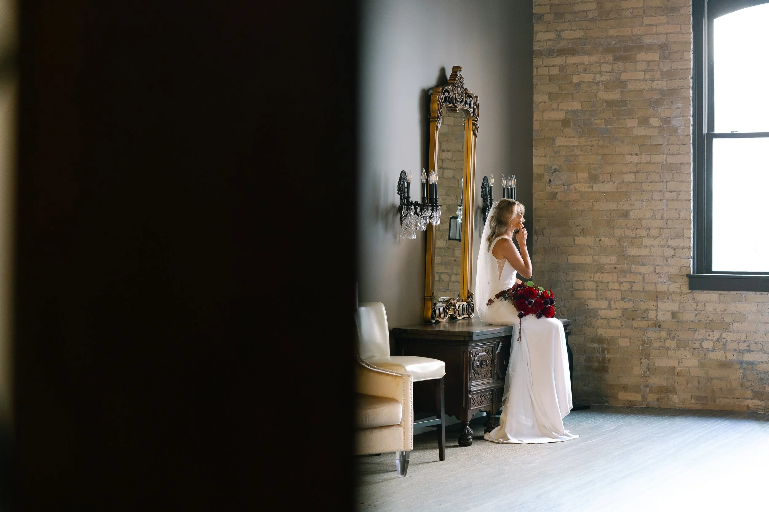 A woman in a white dress sitting on a vintage wooden table, holding a bouquet of red and dark flowers, in a room with a large window, exposed brick wall, and decorative mirror with black sconces.