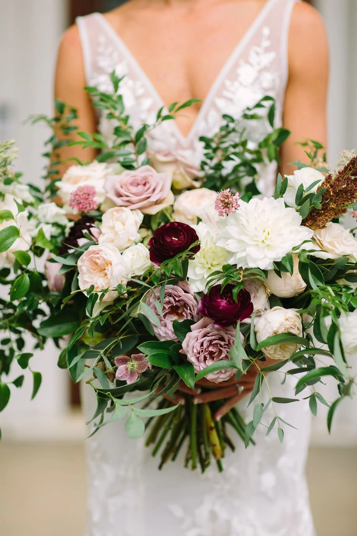A woman in a white lace dress holding a large bouquet of mixed pink, cream, red, and white flowers with greenery.