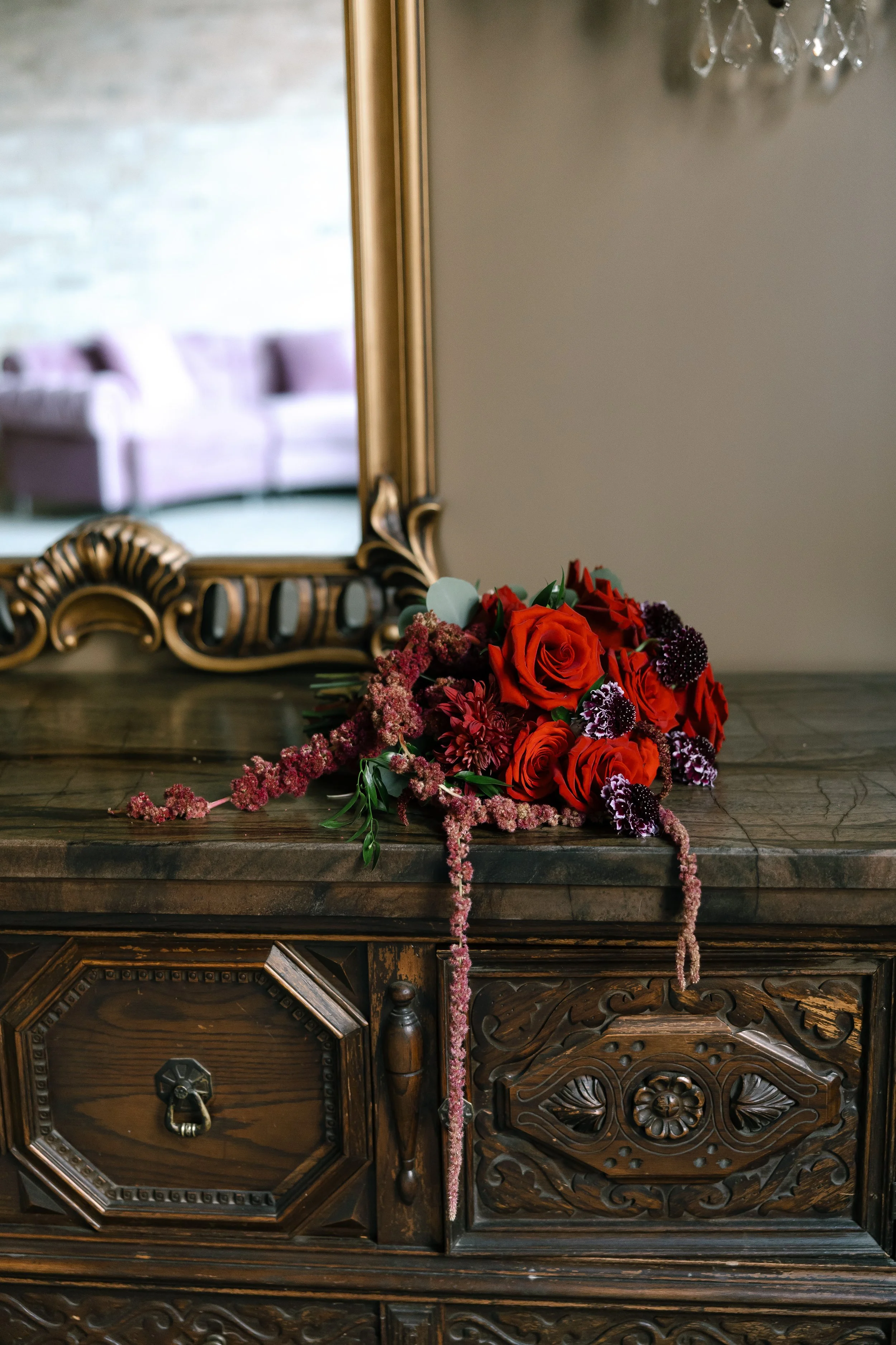 A bouquet of red, purple, and pink flowers resting on a dark wood carved table, with a large ornate mirror reflecting a purple sofa and a brick wall in the background.
