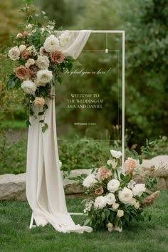 A wedding welcome sign on a transparent board, decorated with a floral arrangement of roses, placed outdoors with green trees in the background.