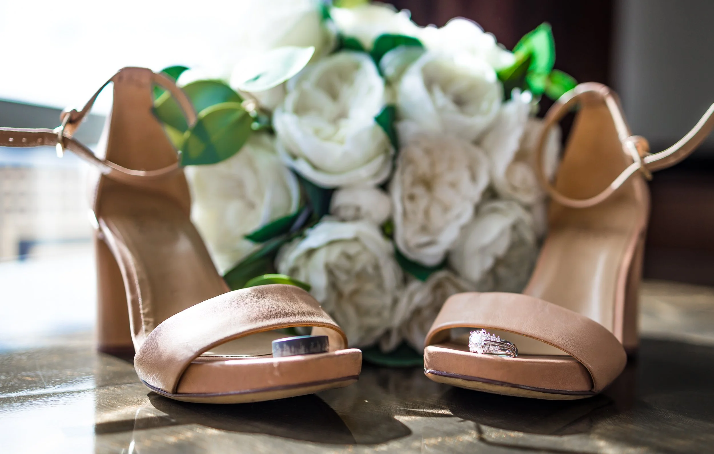 A pair of beige high-heeled shoes on a dark surface with a bouquet of white roses and green leaves in the background.