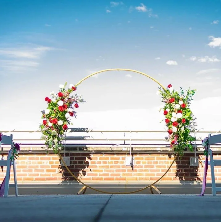 Decorative wedding arch with red, white, and pink flowers on a rooftop with a brick wall and blue sky in the background.