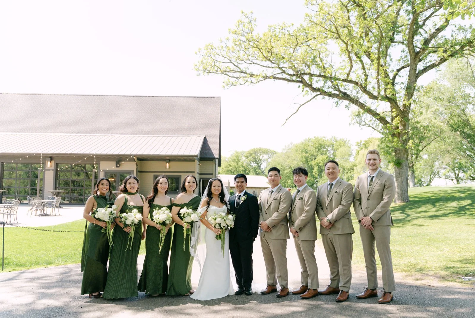 Wedding party standing outside on a sunny day, with bridesmaids in dark green dresses holding white bouquets, and groomsmen in beige suits, with a bride and groom in the center, near a large tree and a building with string lights.