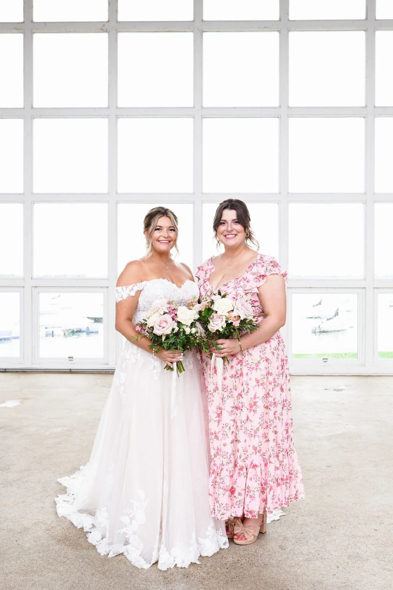 Two women, one in a white wedding dress and the other in a pink floral dress, standing side by side indoors in front of large window panes, both smiling and holding bouquets of flowers.