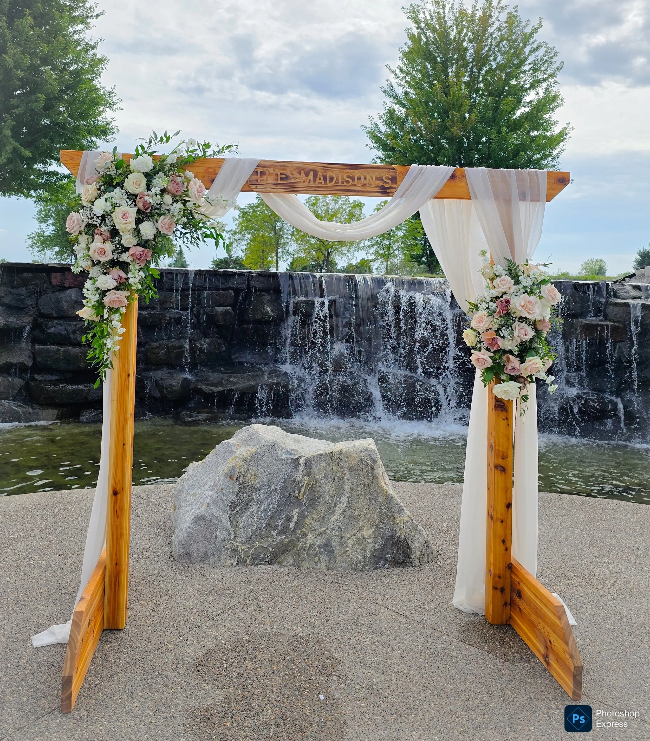 A wooden wedding arch decorated with white and pink roses, white lilies, and greenery, draped with white and sheer curtains, with a sign reading 'The Madison's' at the top, set up outdoors over a waterfall feature with a stone wall, trees, and a part