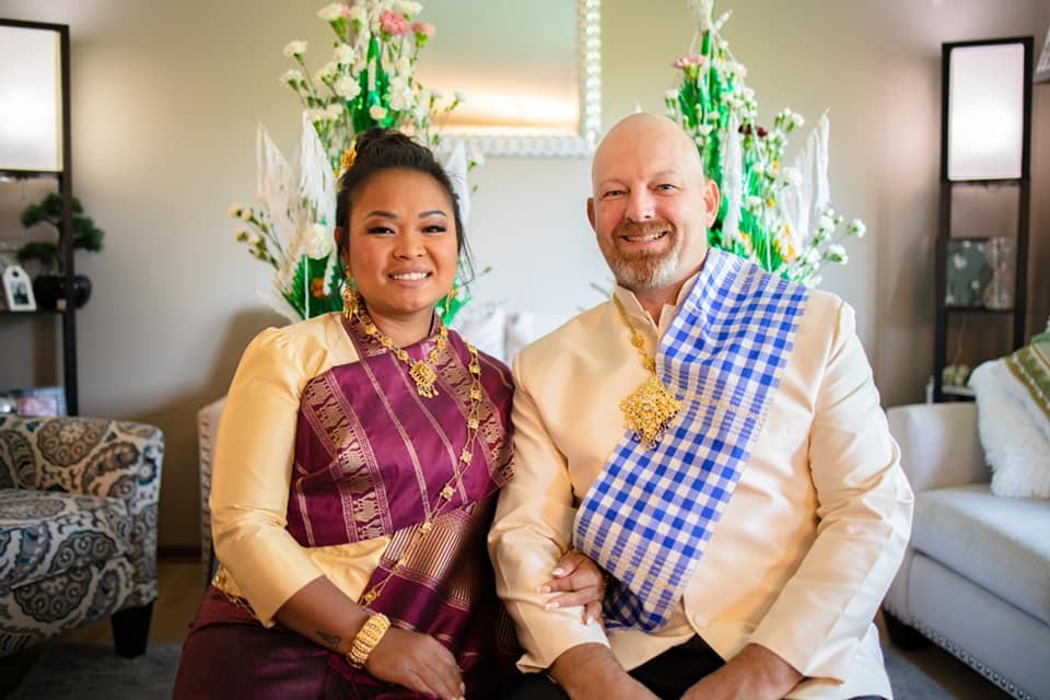 A couple dressed in traditional attire sitting together and smiling, with floral decorations and a mirror in the background.