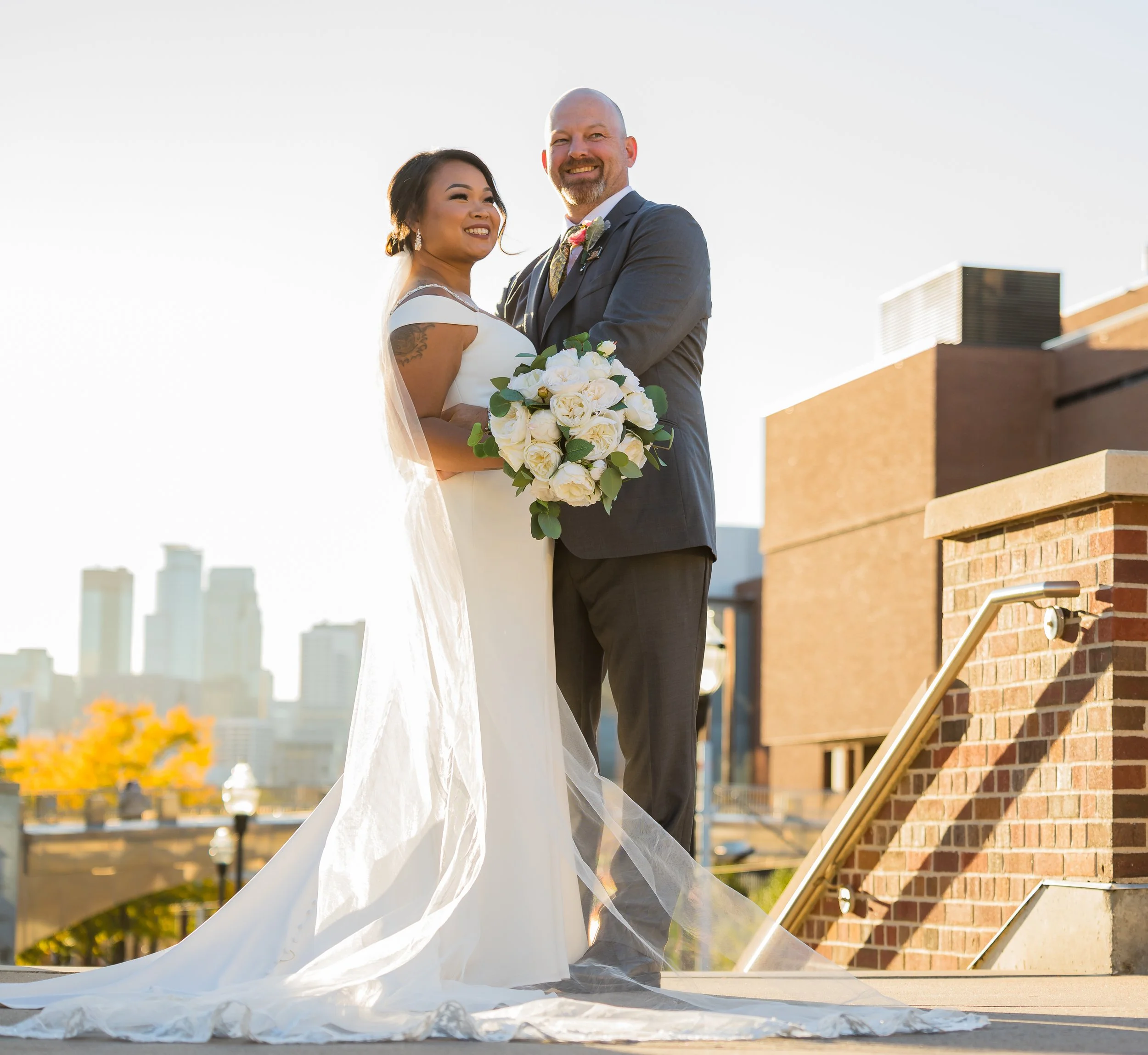A newlywed couple standing on a rooftop terrace, smiling and posing for a photo with city buildings in the background during daylight, with the bride holding a bouquet of white flowers.
