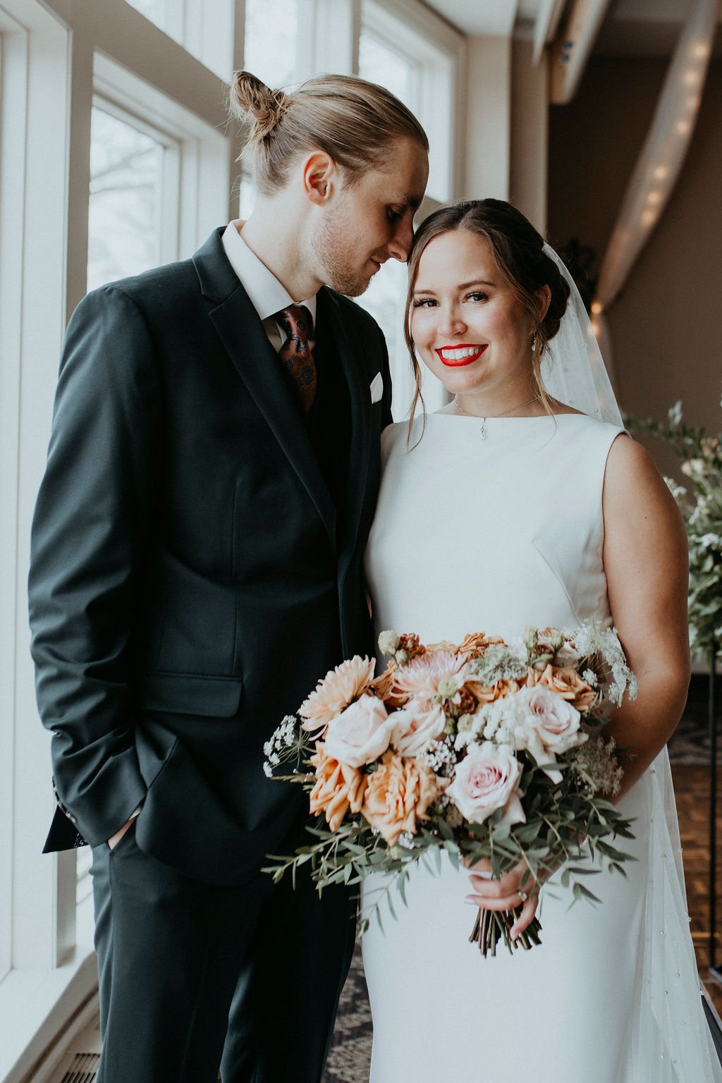 A newlywed couple stands close together indoors near windows, with the bride smiling and holding a large bouquet of pink, peach, and white flowers. The groom, dressed in a black suit and tie, leans his forehead gently against the bride's temple.