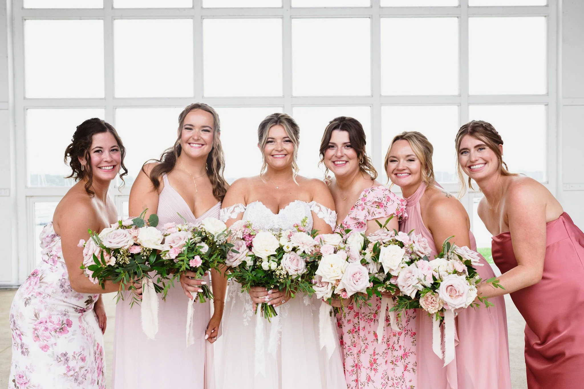 A bride and six women, likely bridesmaids, smiling and posing together in front of large windows indoors, each holding a bouquet of flowers, all dressed in pastel-colored dresses with pinks, creams, and floral patterns.