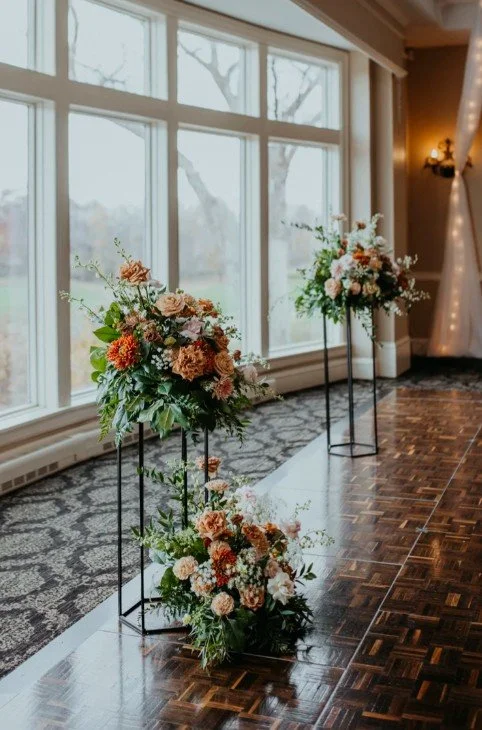 Two tall floral arrangements with pink, white, and peach flowers and greenery on black metal stands, positioned indoors next to large windows in a decorated event space.