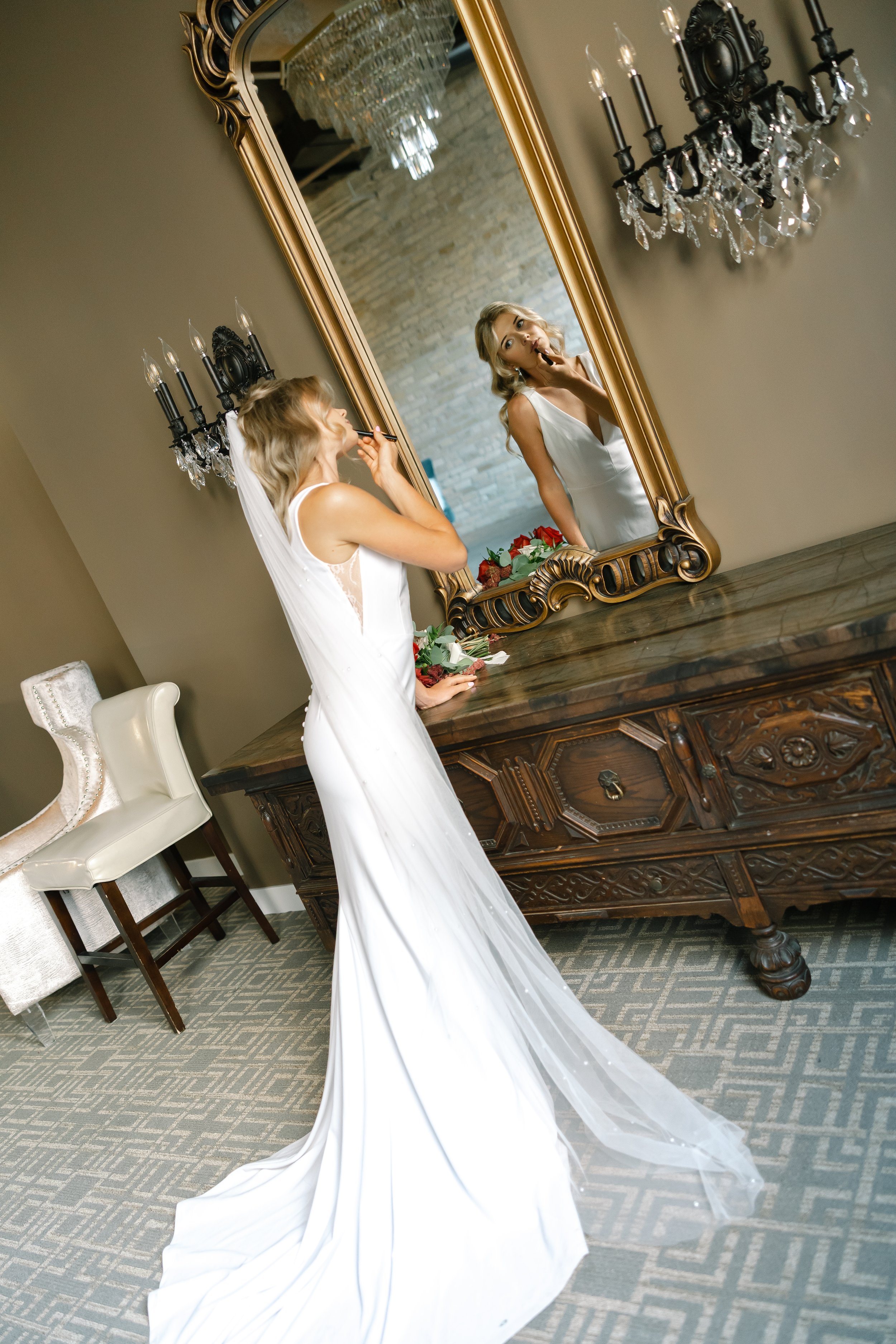 A woman in a white wedding dress doing makeup in front of a large ornate mirror, with crystal chandeliers and vintage furniture in the background.