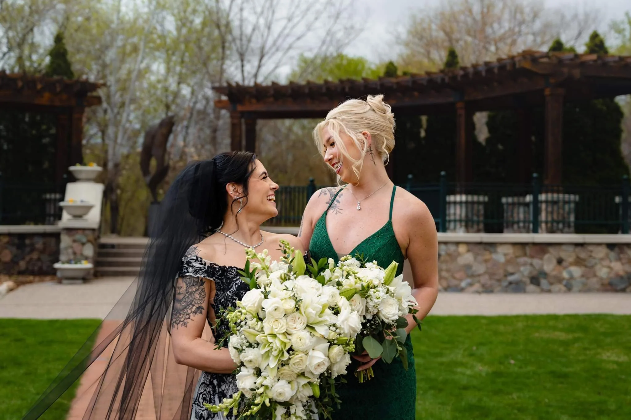 Two women at a wedding, one in a black lace dress with a veil and the other in a green dress holding a large bouquet of white flowers, smiling at each other outdoors with trees and a wooden pergola in the background.