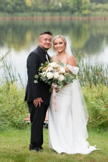A newlywed couple standing by a lake, the bride in a white wedding gown holding a bouquet, and the groom in a black suit, smiling with nature in the background.