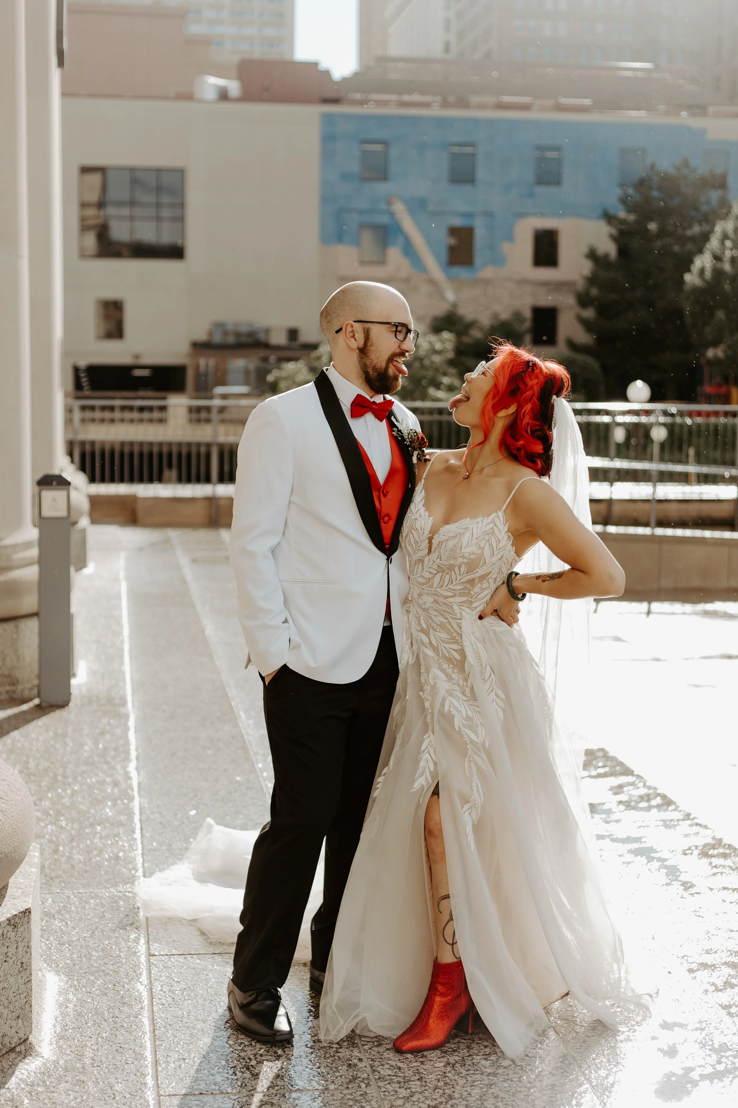A couple in wedding attire standing outdoors on a sunny day in an urban environment, looking at each other and sticking out their tongues playfully.