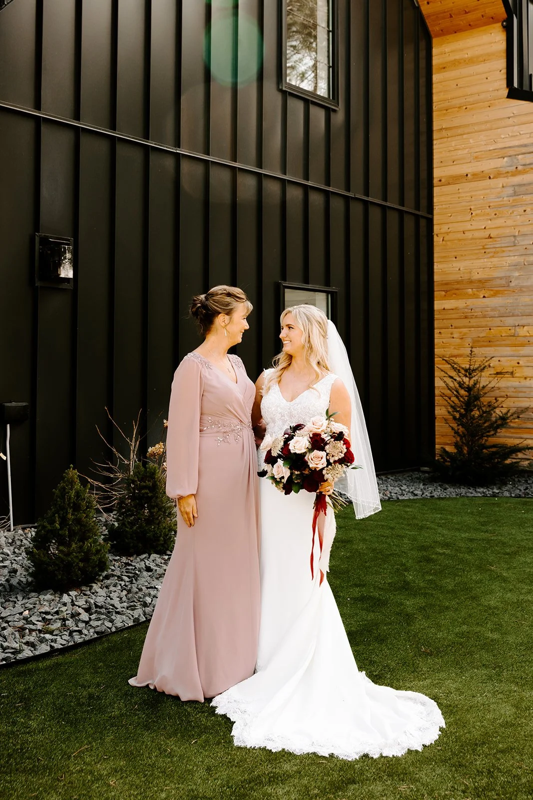 A bride and woman in a mauve dress sharing a moment outdoors beside a modern black and wood building, with small trees and grass in the background.