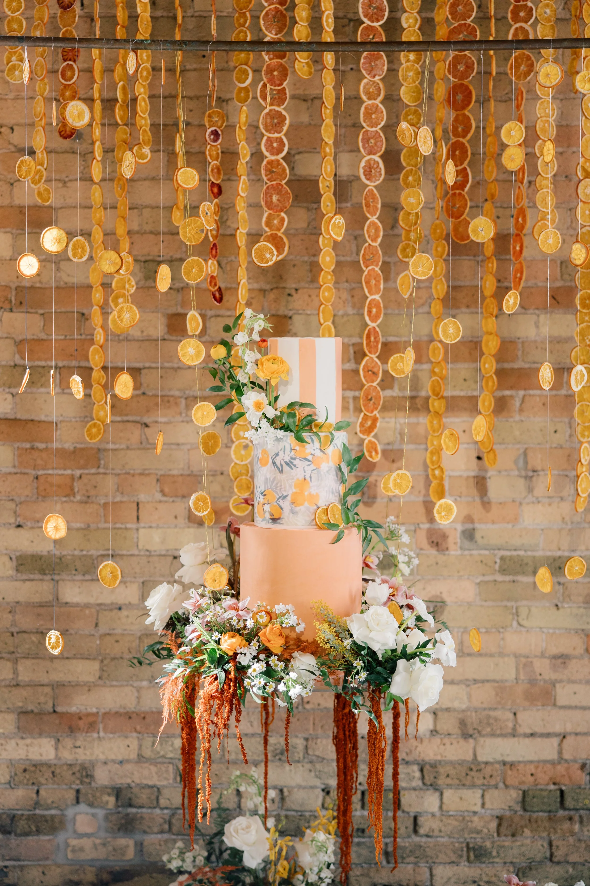 A tiered peach-colored cake decorated with white and orange flowers and green leaves, surrounded by a floral arrangement and hanging dried citrus slices on strings, set against an exposed brick wall.