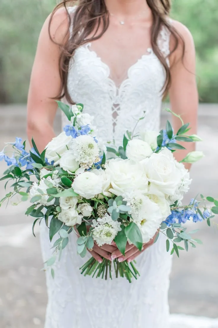 Bride holding a large bouquet of white and blue flowers with green foliage, wearing a white lace wedding dress with a V-neckline and lace details.