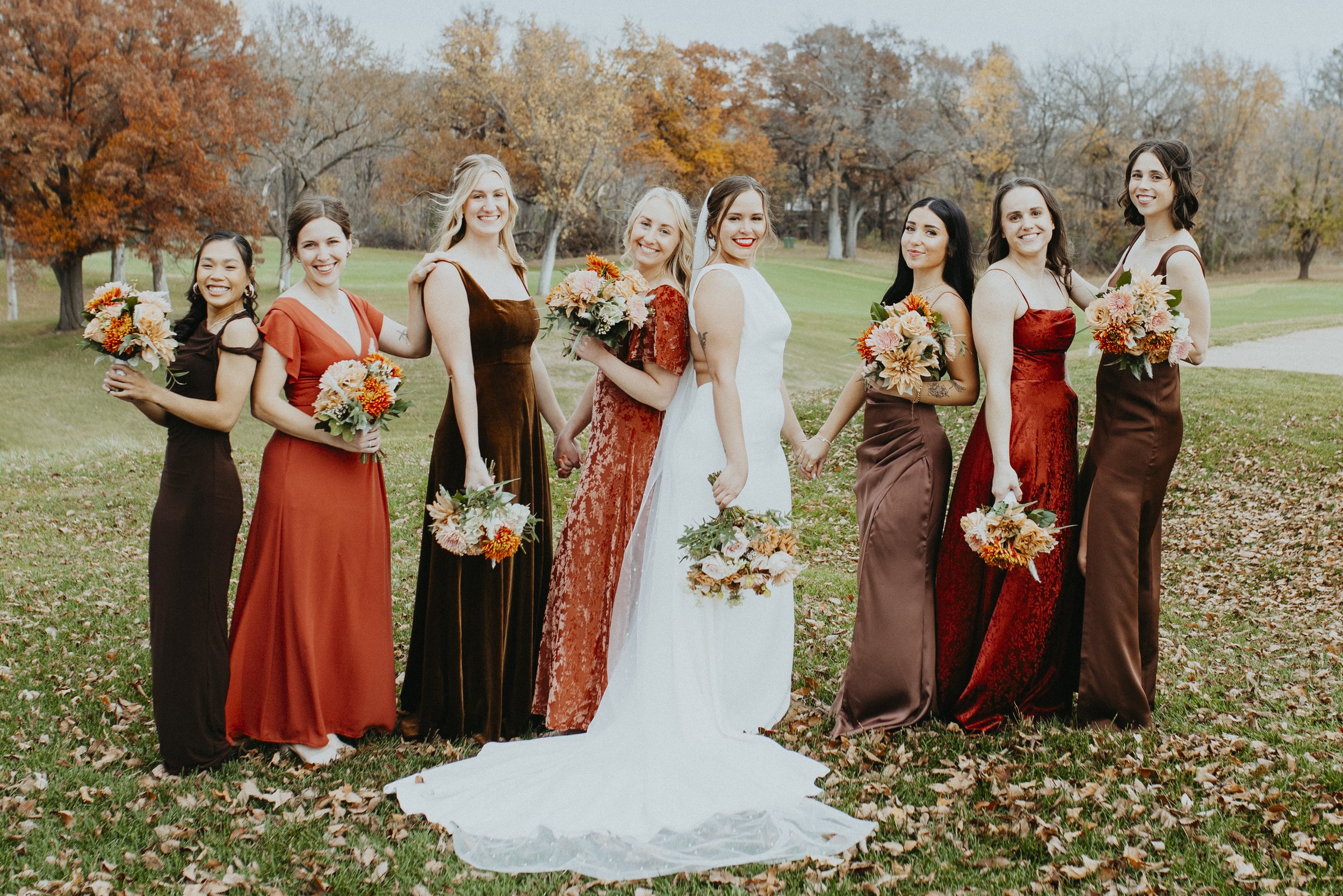 A group of nine women standing outside on a grassy field with autumn trees in the background, holding bouquets of flowers, dressed in formal dresses, smiling, and posing for a photo, with the bride in a white wedding gown in the center.