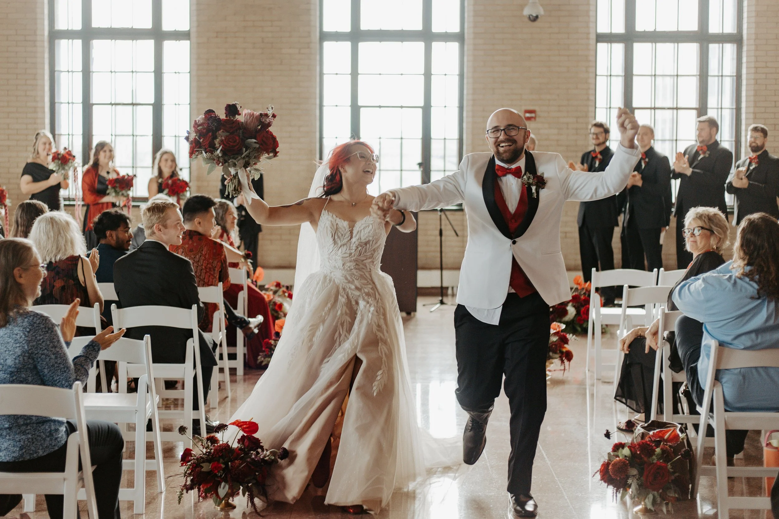A bride and groom happily dancing in the center of a wedding reception hall with large windows, surrounded by seated guests and bridesmaids holding bouquets, all smiling and clapping.