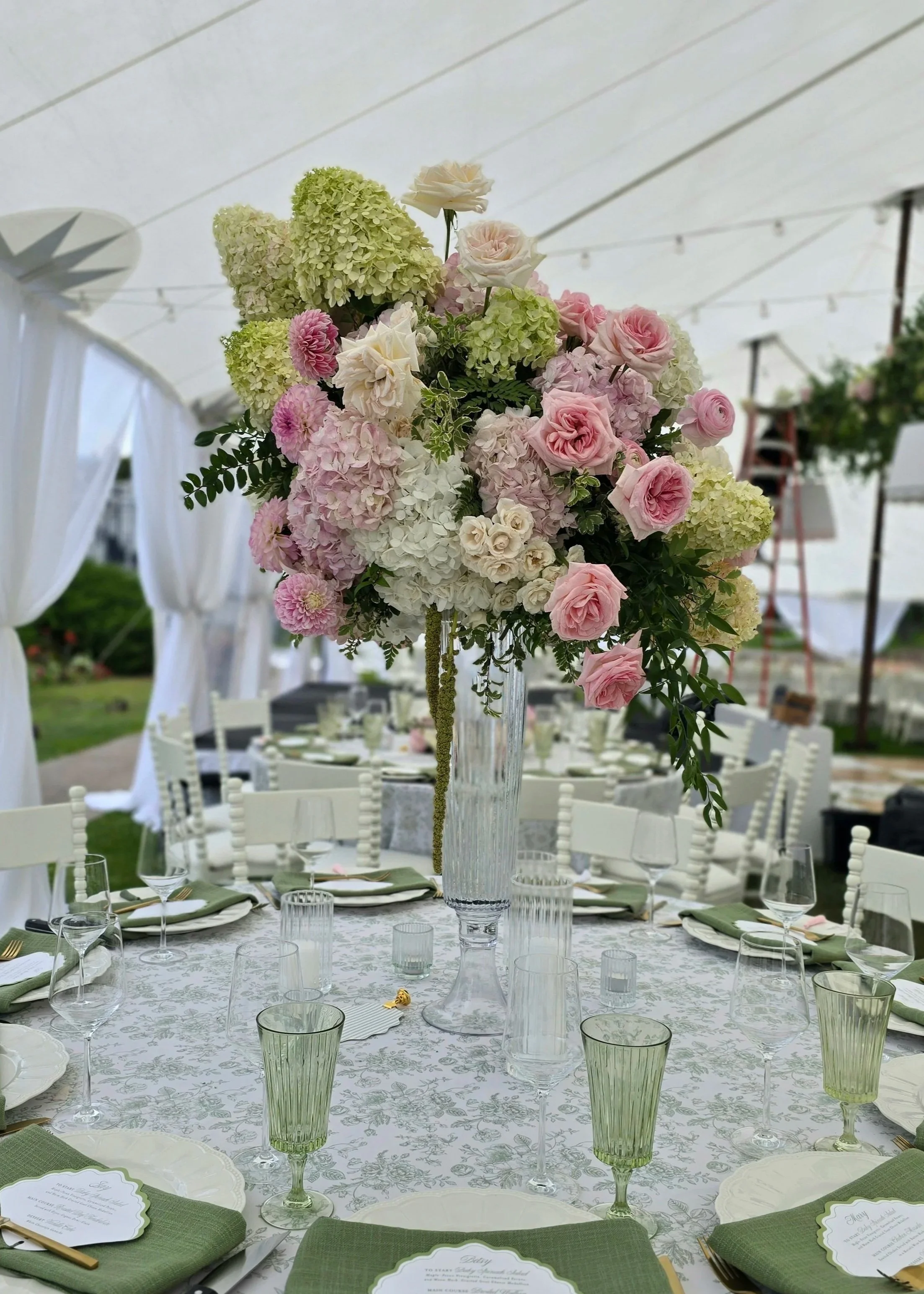 A tall flower arrangement with pink roses, white hydrangeas, and green foliage in a glass vase on a decorated table, set for a formal event under a white canopy with white drapes and outdoor lighting.