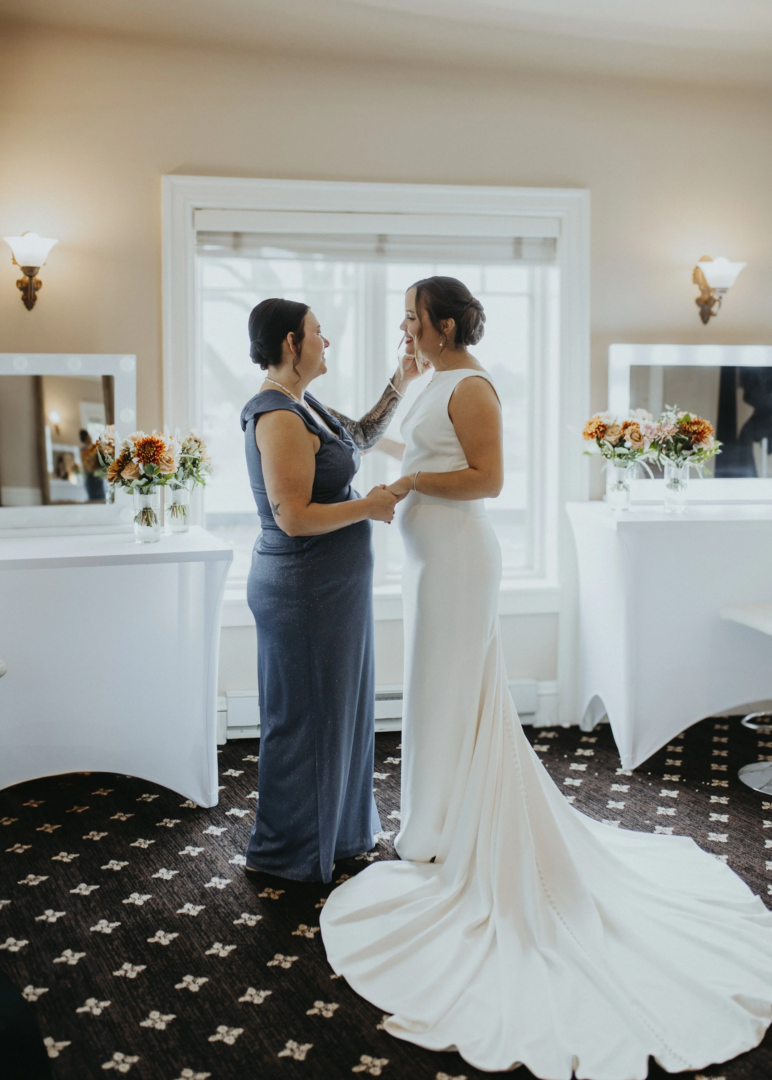Two women stand facing each other, holding hands, in a room with cream-colored walls and floral carpet. One woman is wearing a white wedding dress, and the other is dressed in a dark gown. They are smiling and touching each other's faces in an intimate moment, possibly during a wedding or commitment ceremony, with floral arrangements and mirrors in the background.