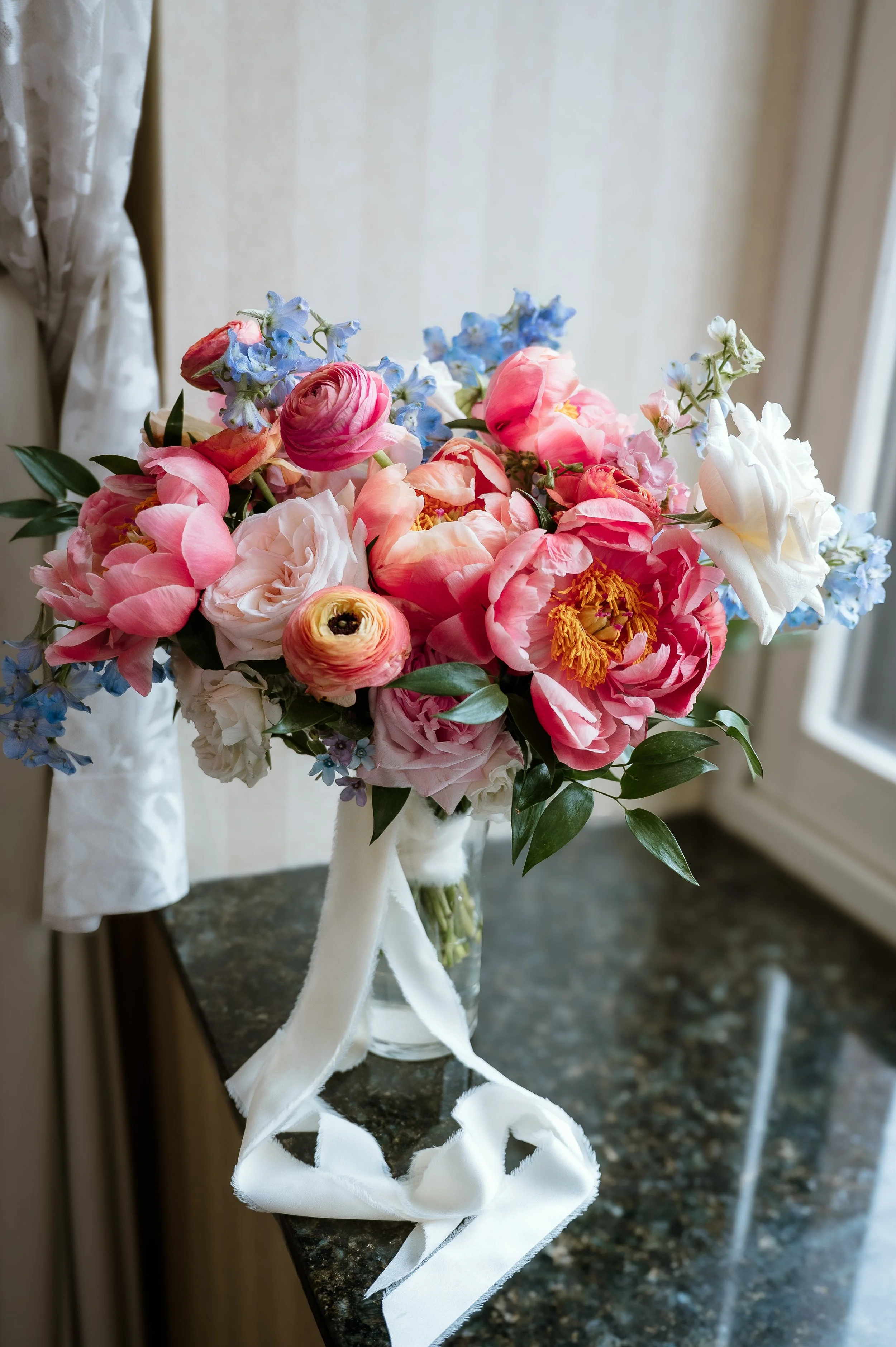 A bouquet of pink, white, and blue flowers with green leaves in a clear glass vase, tied with a white satin ribbon, on a dark granite countertop near a window.