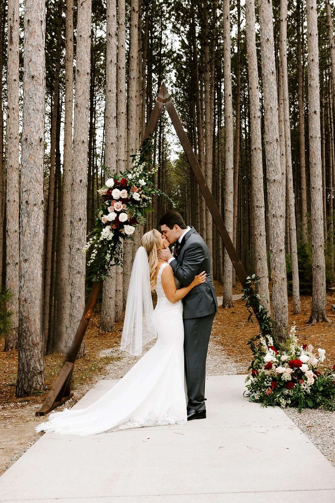A bride and groom sharing a kiss at their outdoor wedding ceremony in a forest, standing on a white rug beneath a triangular floral arch decorated with white and red flowers and green foliage.