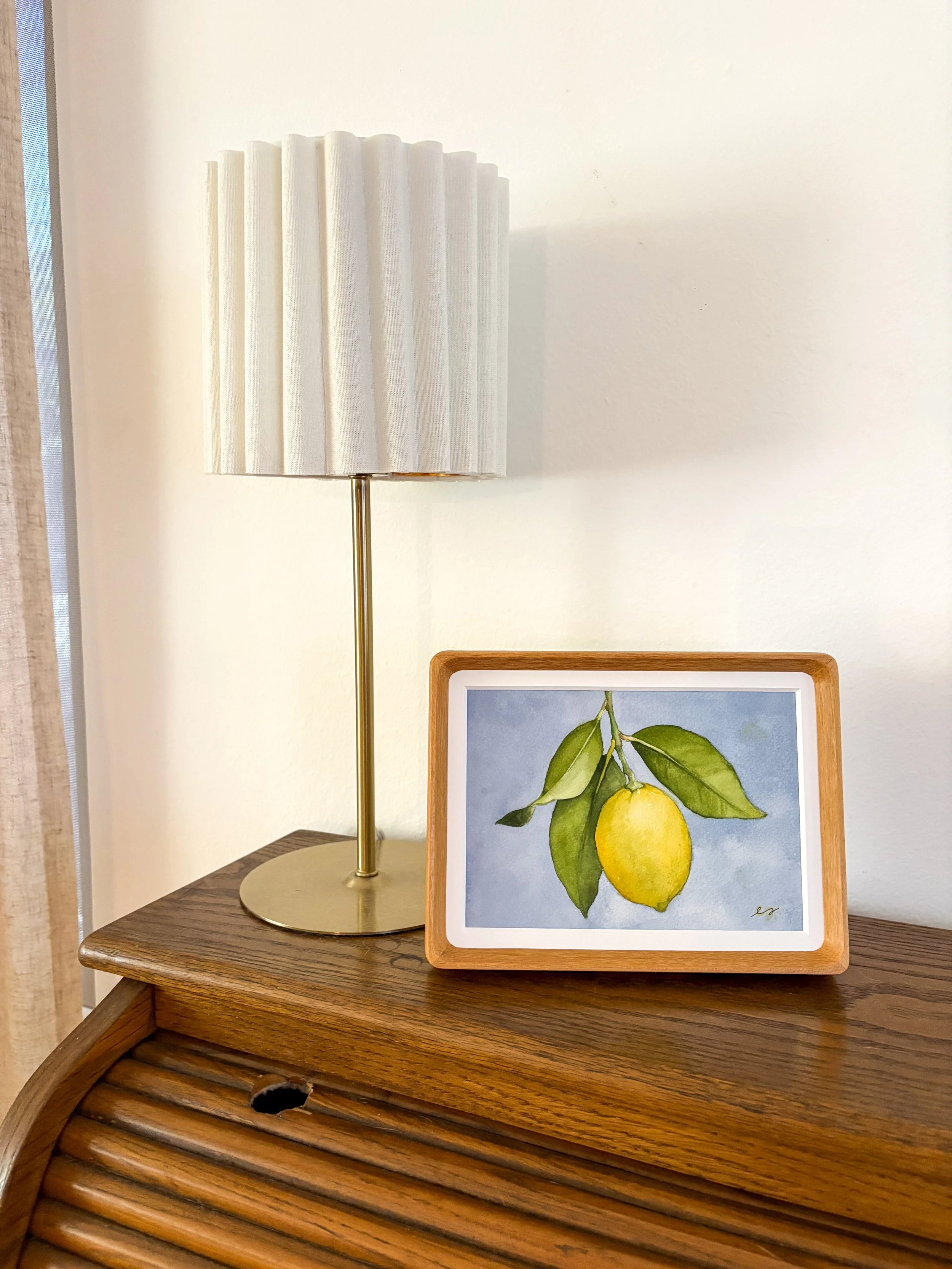 Original small framed watercolor painting of a single yellow lemon on a branch with green leaves against a soft periwinkle blue background, in a warm wood frame