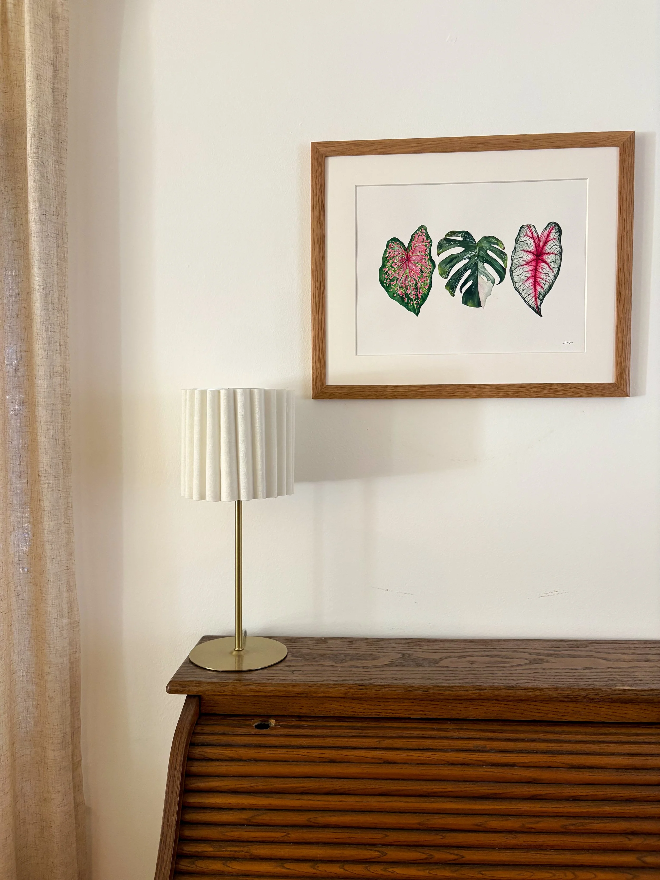 Original framed watercolor painting of three tropical leaves side by side — a pink caladium, variegated monstera, and silver caladium — in a warm wood frame