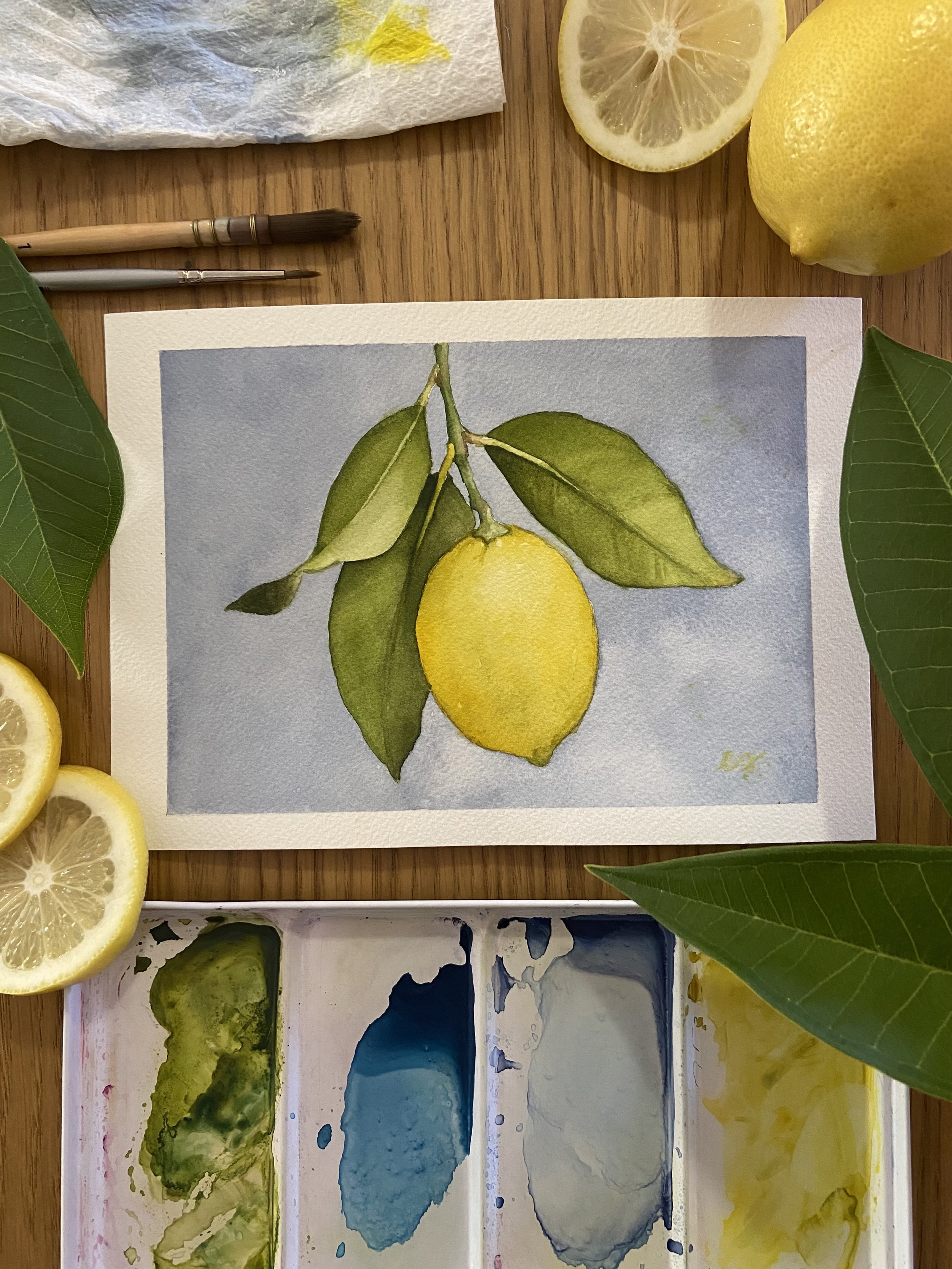 Original small framed watercolor painting of a single yellow lemon on a branch with green leaves against a soft periwinkle blue background, in a warm wood frame