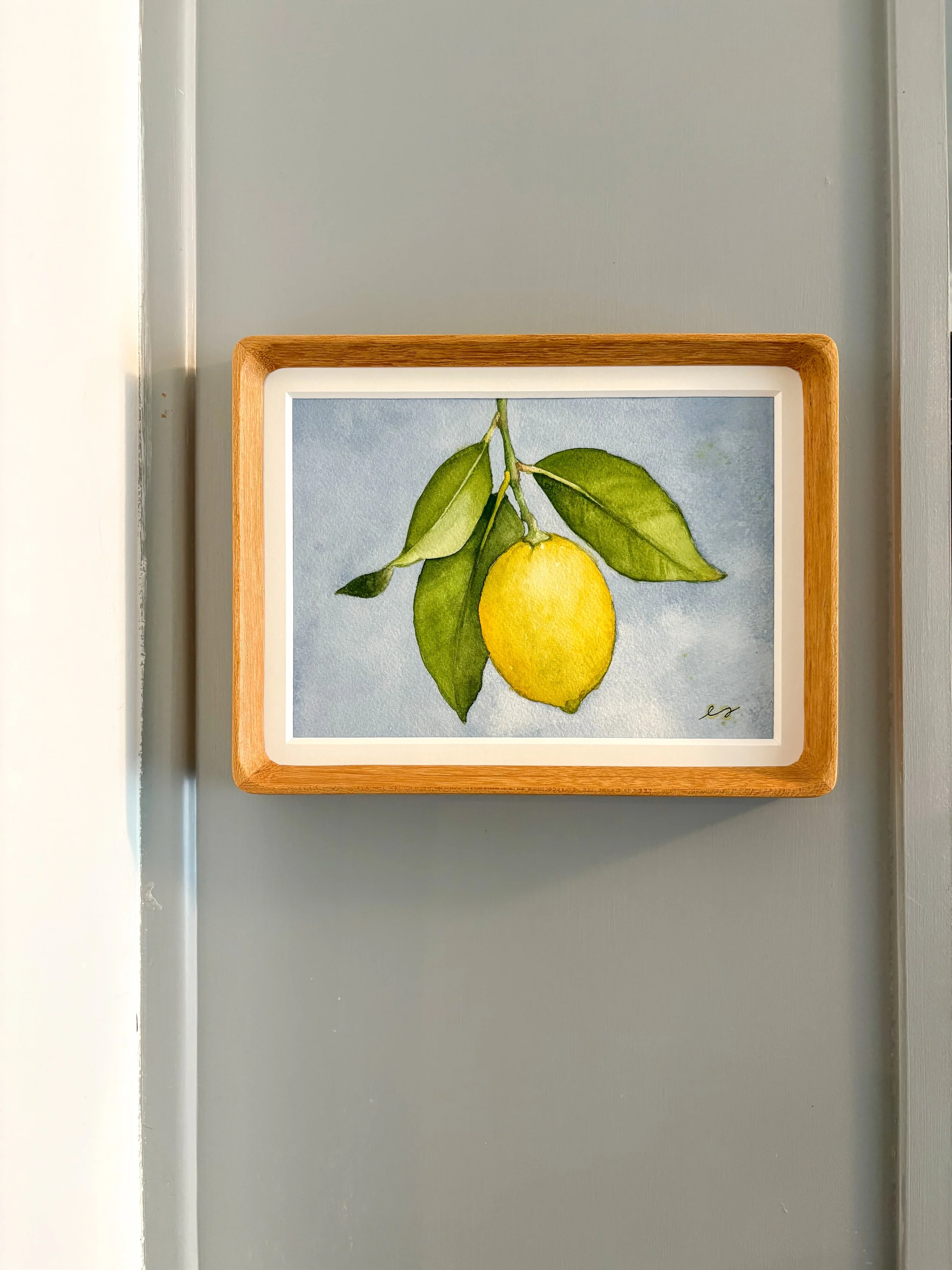 Original small framed watercolor painting of a single yellow lemon on a branch with green leaves against a soft periwinkle blue background, in a warm wood frame