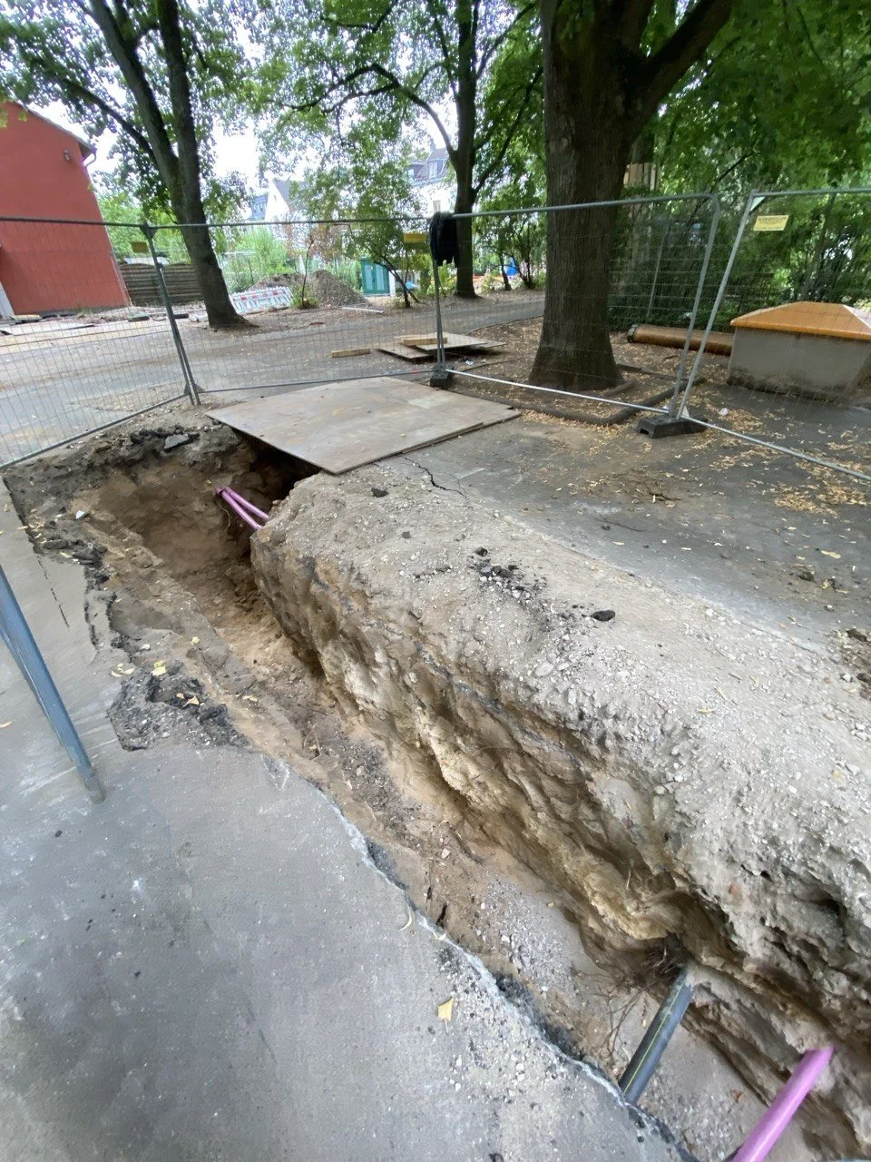 Eine Baugrube auf einer Gehwegseite mit mehreren pinkfarbenen Rohren, umgeben von Bauzäunen und Bäumen, in einer städtischen Umgebung.