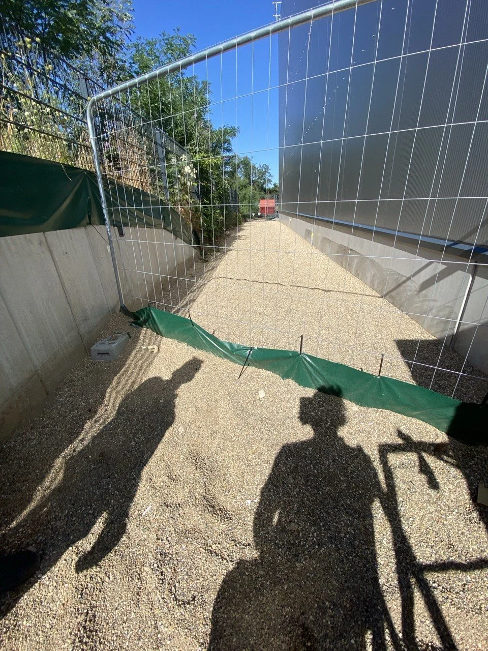 Ein Bauzaun mit grünem Schutznetz auf einem Kiesweg, Schatten von zwei Personen, Fahrradschatten, und blauer Himmel im Hintergrund.