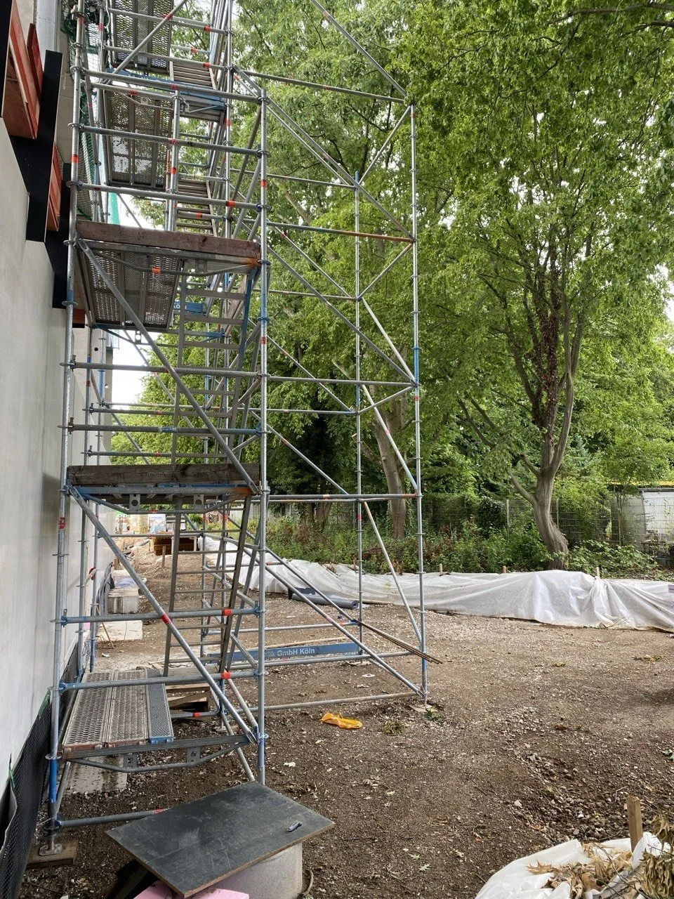 Baustelle mit Metallgerüst neben einem weißen Gebäude, im Hintergrund Bäume und eine weiße Schutzplane auf dem Boden.