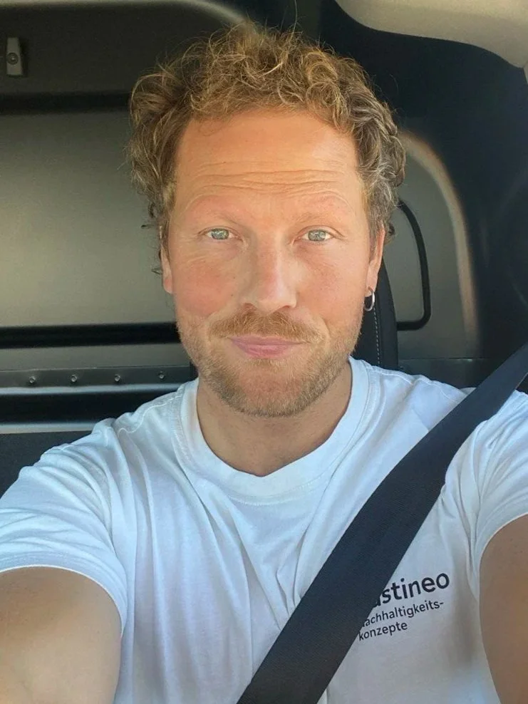 Ein Mann mit kurzen, lockigen, blonden Haaren und einem leichten Bart trägt ein weißes T-Shirt und sitzt im Auto.